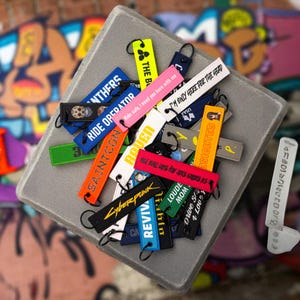 May include: A collection of colorful keychains with various text and graphic designs. The keychains are in various colors, including black, blue, orange, pink, and green. Some keychains have text like "Cyberpunk" and "Ride Operator".