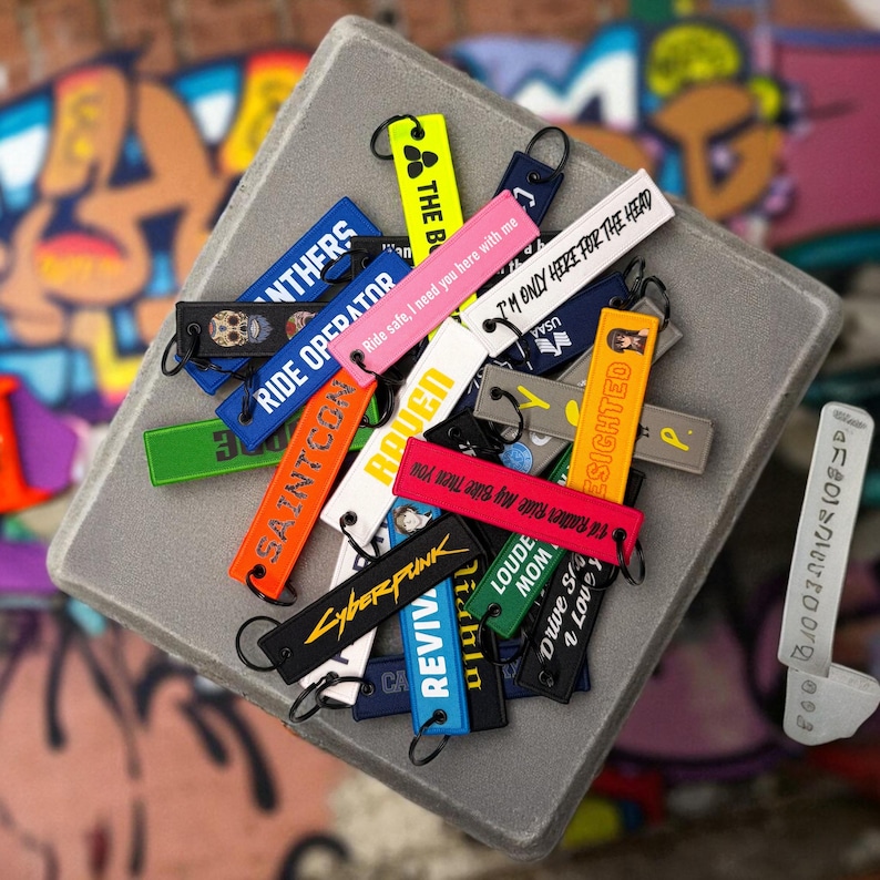 May include: A collection of colorful keychains with various text and graphic designs. The keychains are in various colors, including black, blue, orange, pink, and green. Some keychains have text like "Cyberpunk" and "Ride Operator".