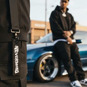 May include: Black keychain with the text "NEVERMIND" in white. The keychain is attached to a black garment. A person in a black hoodie and pants is leaning against a blue car.