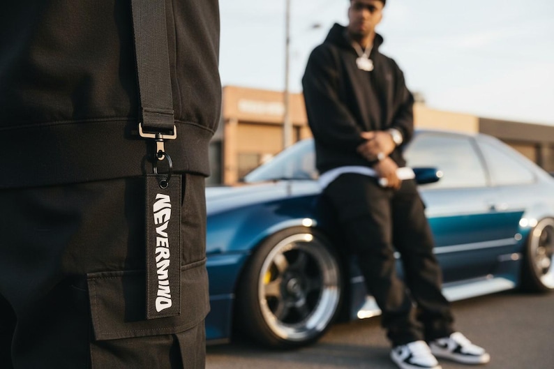 May include: Black keychain with the text "NEVERMIND" in white. The keychain is attached to a black garment. A person in a black hoodie and pants is leaning against a blue car.
