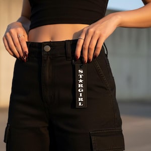 May include: Black cargo pants with a matching black tag that reads "STARGIRL" in white capital letters. The tag is hanging from the pants' belt loop. The image is taken in natural light.