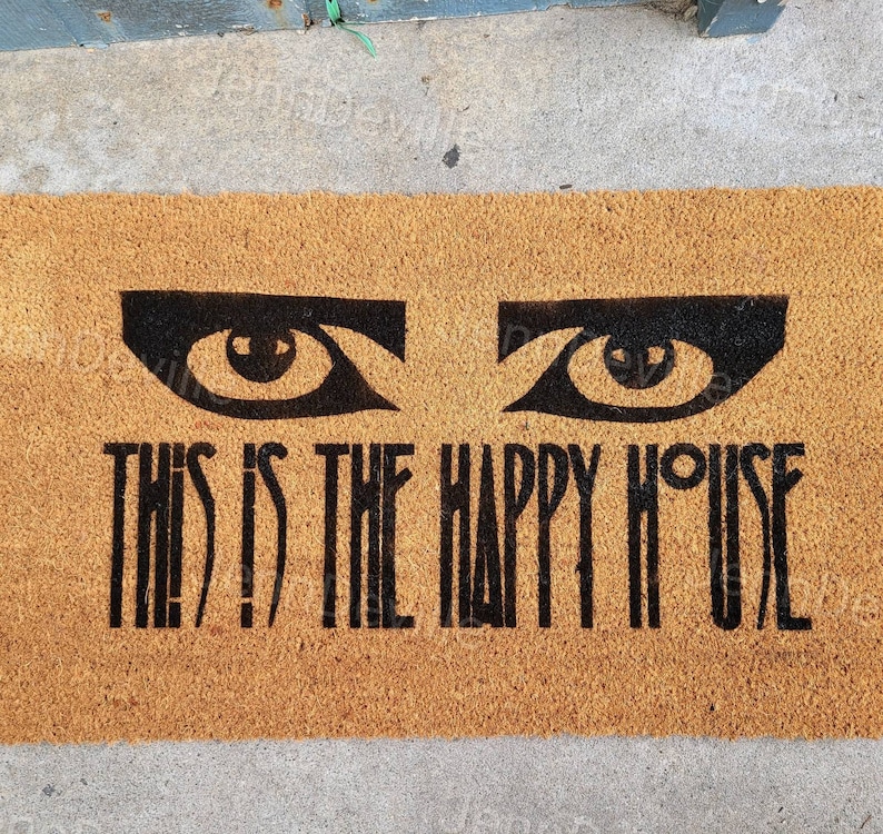 Puede incluir: Una alfombra de coco marr&oacute;n con el texto "This is the Happy House" en letras negras. Dos ojos negros con pupilas blancas est&aacute;n por encima del texto.