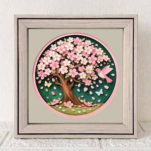 Puede incluir: Obra de arte enmarcada que representa un cerezo en flor estilizado con flores rosas, un tronco marrón y un pájaro rosa. La obra está sobre un fondo verde con pétalos y mariposas cayendo, enmarcada en madera clara.
