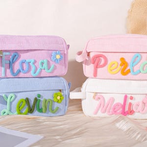 Op de afbeelding: Vier corduroy make-up tassen in pastelkleuren: lavendel, roze, lichtblauw en wit. Elke tas heeft een naam geborduurd in kleurrijk garen, met een bloemaccent. De tassen hebben een ritssluiting en een klein handvat.