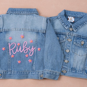 May include: Two light blue denim jackets for children. One jacket has the name "Ruby" embroidered in pink with pink flower accents on the back. The other jacket is plain with button closures and a small tag.