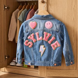 May include: A blue denim jacket with pink patches, including a heart, smiley face, and lightning bolt. The name "SILVIE" is spelled out in pink letters on the back. The jacket is hanging in a wooden wardrobe with other clothing.