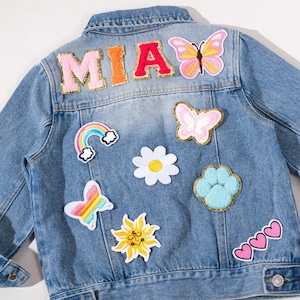 Pode incluir: Jaqueta jeans azul claro decorada com patches bordados coloridos. Os patches incluem um arco-íris, borboletas, uma margarida, um sol, uma pegada, corações e o nome "MIA".