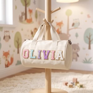 May include: A cream-colored cylindrical duffel bag featuring the name OLIVIA in large, multi-colored, textured varsity-style letters across the front. The bag has two top handles and is suspended from a wooden stand by its straps.