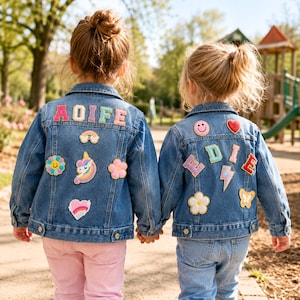 May include: Two children wearing blue denim jackets adorned with colourful patches and name patches. One jacket says "AOIFE" and the other says "EDIE". The jackets also feature patches of a rainbow, flowers, a unicorn, a heart, a smiley face, and a lightning bolt.