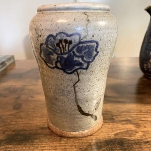 May include: A ceramic vase with a light beige speckled finish. A large blue floral design is painted on the front. The vase has a rounded top and a slightly flared base. The vase is approximately 6 inches tall.
