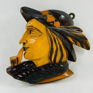 May include: A carved wooden wall hanging depicting a man in profile, smoking a pipe. The figure has a yellow face and hair, a black hat with orange accents, and a black scarf. The pipe is also yellow. A small brass ring is at the top.