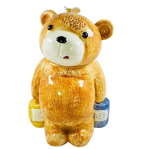 Puede incluir: Un tarro de galletas de cerámica con forma de oso y acabado brillante. El oso es de color marrón claro con una cara blanca, ojos negros y una boca roja pequeña. Sostiene dos pequeños botes de miel, uno amarillo y otro azul, con la palabra "HONEY". Una pequeña abeja está encima.