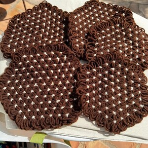 May include: Four brown and white crocheted coasters. Each coaster is square with a scalloped edge and a repeating pattern of white star-shaped designs. The coasters are made of a thick yarn and appear handmade.