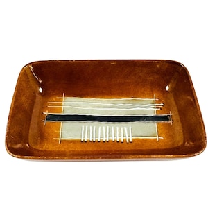 Puede incluir: Plato rectangular de cerámica marrón con un diseño moderno de mediados de siglo. El plato presenta un diseño central de líneas horizontales en negro, blanco y gris, con líneas blancas verticales en la parte inferior. El plato tiene esquinas redondeadas.