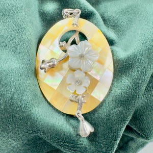 以下が含まれることがあります： 黄色と白のチェック柄の楕円形ペンダント。白い花のアクセントと銀色の金属のディテールで飾られています。ペンダントには吊り下げ用のベイルが付いており、おそらくジュエリーです。