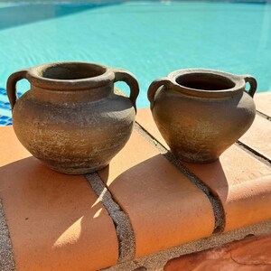 May include: Two small, rustic brown ceramic pots with handles. The pots have a rounded shape and a textured surface, sitting on a terracotta ledge with a pool in the background. The pots are likely for decorative purposes.
