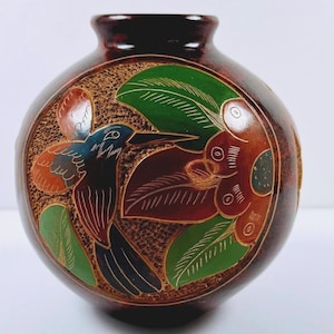 Op de afbeelding: Een ronde, donkerbruine vaas met een getextureerd oppervlak en een gegraveerd ontwerp van een kolibrie in vlucht bij een grote bloem. De vogel- en bloemelementen zijn ingevuld met levendige groene, oranje en zwarte kleuren voor contrast.