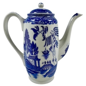 Puede incluir: Una cafetera de cerámica blanca y azul con tapa y boquilla curva. La cafetera presenta un diseño clásico de patrón Willow con escenas de pájaros, árboles y un puente. El asa es curva y está unida al lateral.