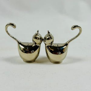 May include: Two silver-colored cat-shaped figurines. Each cat has a curved tail and a pointed ear. The cats are facing each other, with their noses almost touching. The figurines have a shiny, reflective surface and are set against a plain white background.