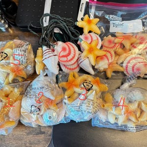 May include: A collection of decorative string lights featuring plastic starfish and seashell covers in shades of orange, pink, and white. The set includes several clear plastic bags containing loose shell and starfish pieces, with one strand of lights partially assembled.