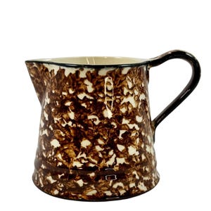 Puede incluir: Una jarra de cerámica moteada marrón y blanca con asa y borde negros. La jarra tiene un pico para verter y un interior color crema. El patrón moteado le da a la jarra un aspecto rústico y vintage. Un artículo de cocina clásico.