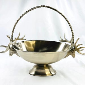 Può includere: Una ciotola in metallo color oro con base a piedistallo e manico intrecciato. La ciotola presenta due decorazioni a forma di testa di cervo color argento con corna sui lati opposti, dove il manico si collega al bordo, creando un recipiente decorativo.