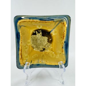 May include: Square ceramic dish with a yellow interior and a dark blue rim. The center of the dish features a dark brown circle with a cluster of crystal-like formations. The dish is displayed on a clear plastic stand.