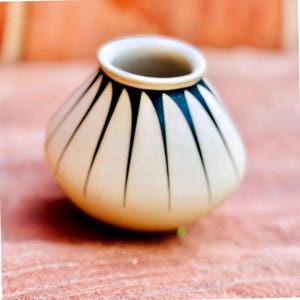 Puede incluir: Pequeño jarrón de cerámica color crema, cuerpo redondeado y abertura ancha. Decorado con formas triangulares negras alargadas que irradian desde el borde. El borde es blanco, y está sobre una superficie texturizada, rojo-anaranjada.