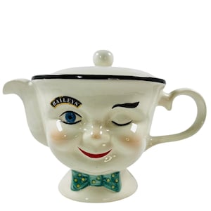 May include: A cream-colored ceramic teapot with a face design, featuring a winking eye, rosy cheeks, and a red smiling mouth. The teapot has a black rim, a green bow tie, and the word "BAILEY'S" on the side. It has a lid with a small white knob.