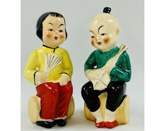 Könnte beinhalten: Zwei Vintage-Keramikfiguren, die asiatische Figuren darstellen. Eine Figur trägt eine gelbe Jacke und eine rote Hose und hält einen Fächer. Die andere Figur trägt eine grüne Jacke und eine schwarze Hose und hält ein Saiteninstrument. Beide sitzen auf beigen Blöcken.