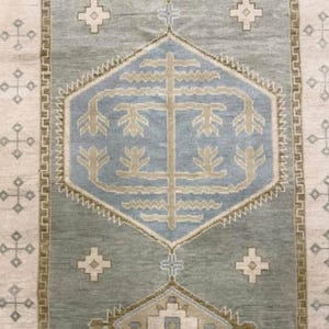 Op de afbeelding: Een rechthoekig tapijt met een centraal geometrisch patroon in blauwe en beige tinten. Het tapijt heeft een lichtgroene achtergrond, een beige rand en decoratieve kruispatronen. Het ligt op een lichtbeige oppervlak.