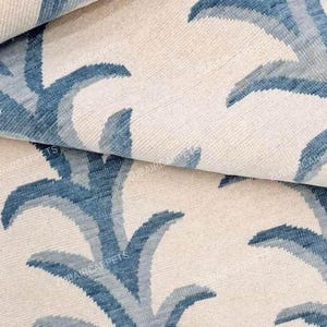 Puede incluir: Primer plano de un textil doblado con un patrón repetido de hojas de palma estilizadas azules y celeste sobre un fondo crema. La tela tiene una textura tejida y el diseño es visible en la superficie.