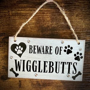 May include: A rectangular wooden sign with the text "BEWARE OF WIGGLEBUTTS" in black. The sign features a black heart with a paw print, paw prints, and bone graphics. The sign is white with a distressed finish and has small, clear rhinestones.