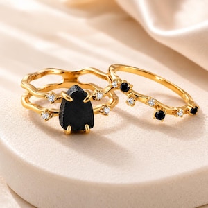 May include: Two gold-toned rings featuring a wavy band design. One ring showcases a central, rough-cut black stone held by prongs, accented with small clear stones. The second ring features a delicate wavy band set with alternating black and clear stones.