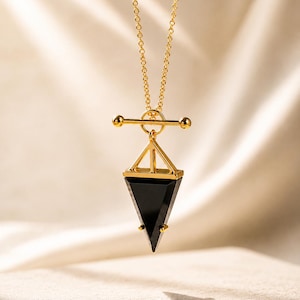 May include: A gold-toned necklace featuring a delicate chain and a toggle clasp. The pendant consists of a geometric gold frame attached to a pointed, triangular black stone, creating a sharp, angular silhouette with a polished finish.