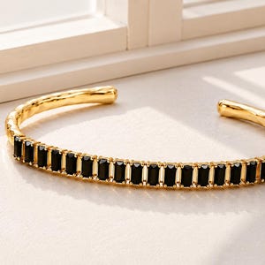 Könnte beinhalten: Ein goldfarbenes Armband in Manschettenform mit einer Reihe rechteckiger schwarzer Steine auf der Vorderseite. Das Metallband hat eine texturierte, gehämmerte Oberfläche und ein offenes Design mit einer Reihe gleichmäßiger, vertikaler schwarzer Edelsteine.