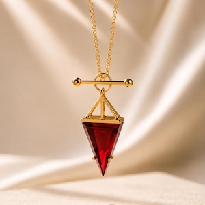 Puede incluir: Un collar de tono dorado con una cadena fina y un cierre de barra. El colgante consiste en una piedra preciosa triangular de color rojo intenso con facetas, engastada en un marco dorado y suspendida de una barra horizontal.