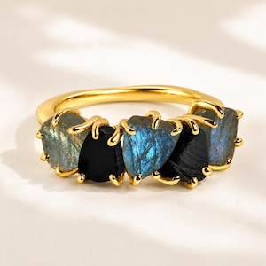 Puede incluir: Un anillo dorado con cinco piedras irregulares y en bruto alternadas. Tres piedras son de labradorita azul grisácea iridiscente con fracturas internas visibles, y dos son negras opacas, sujetas por garras doradas.