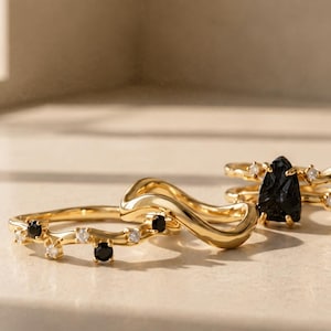 May include: A set of three gold rings. One ring features a wavy band with alternating black and clear stones. A second ring is a plain, smooth, gold wavy band. The third ring has a double band design with a central pear-shaped black stone.