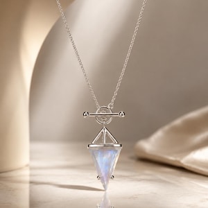 Op de afbeelding: Een zilveren ketting met een knevelsluiting en een driehoekige hanger. De hanger bestaat uit een zilveren frame met daarin een doorschijnende, iriserende steen die een zachte blauwe gloed vertoont. De ketting is gemaakt van fijne schakels.