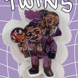 May include: A clear acrylic charm featuring two cartoon characters, one holding a bloody saw and the other a vial. The charm has the word "TWINS" in white outlined letters. The background is a purple grid.