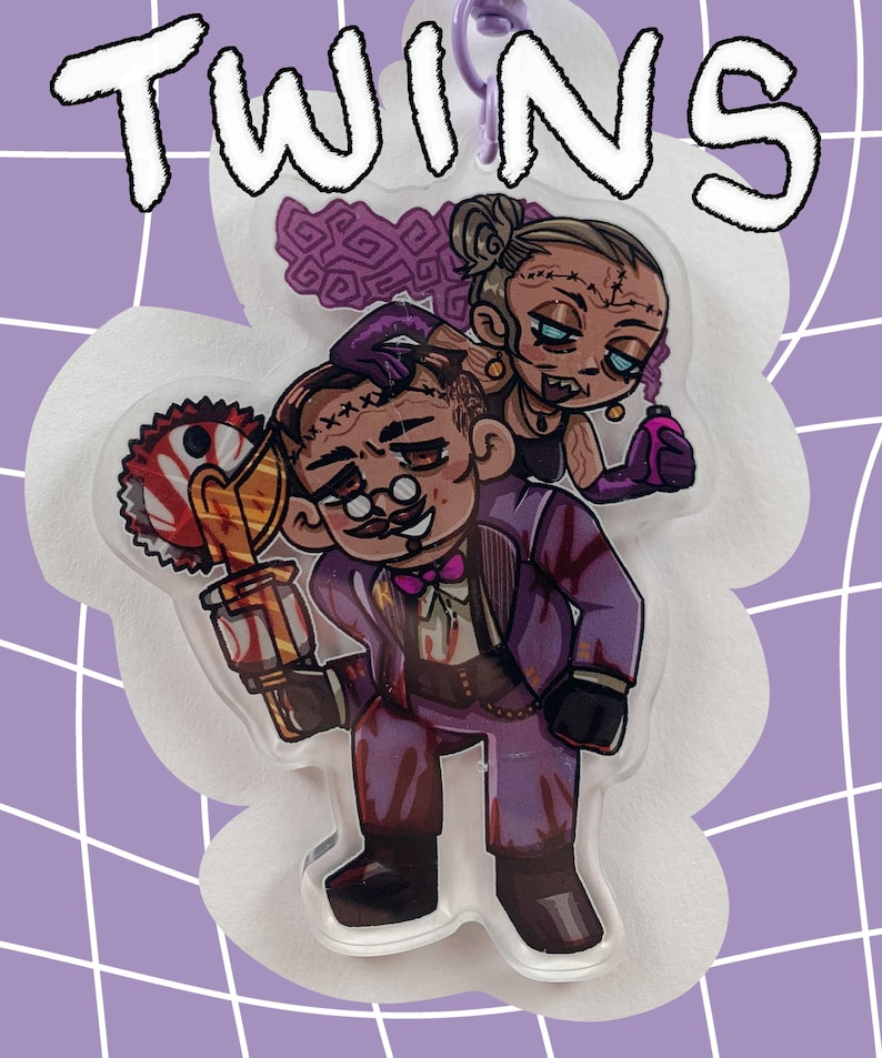 May include: A clear acrylic charm featuring two cartoon characters, one holding a bloody saw and the other a vial. The charm has the word "TWINS" in white outlined letters. The background is a purple grid.