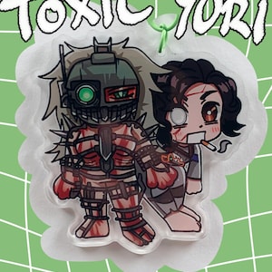 May include: A clear acrylic charm featuring two anime-style characters. One character is in a robotic suit, and the other is holding a cigarette. The charm has the words "TOXIC Yuri" in white, outlined in black, at the top. The background is a light green with a white grid pattern.