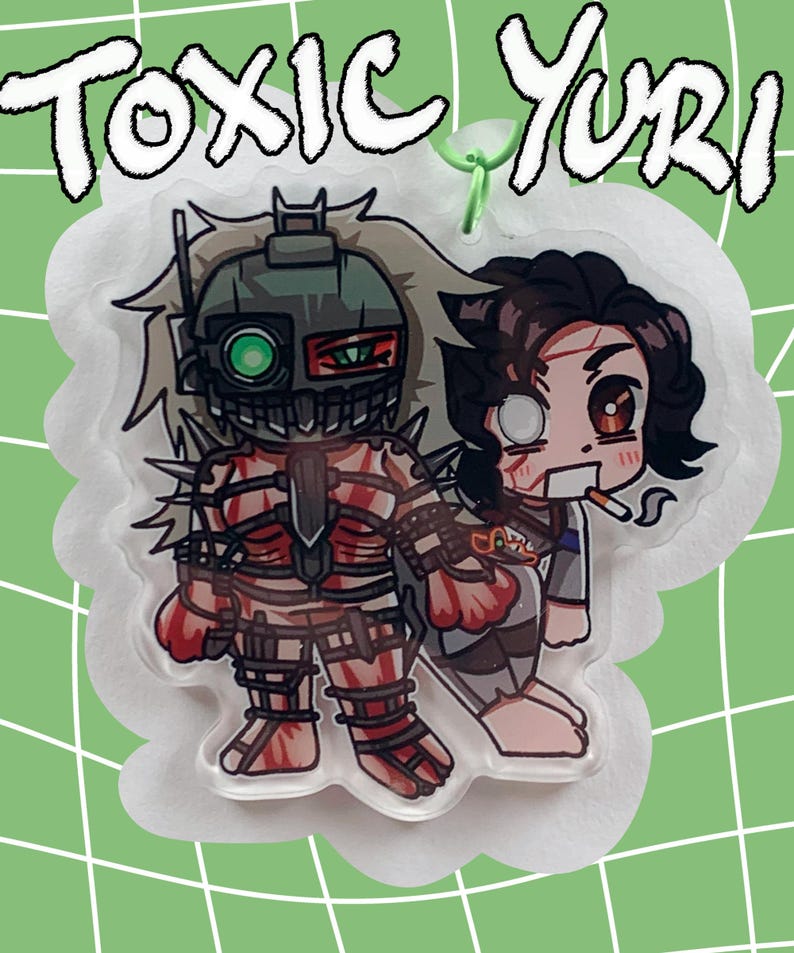 May include: A clear acrylic charm featuring two anime-style characters. One character is in a robotic suit, and the other is holding a cigarette. The charm has the words "TOXIC Yuri" in white, outlined in black, at the top. The background is a light green with a white grid pattern.