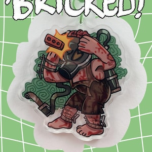 May include: A clear acrylic charm featuring a cartoon figure wearing a gas mask and brown clothing, with blood splatters. The charm has the word "BRICKED!" in white, outlined letters. The charm is on a white, cloud-shaped background, with a green background.