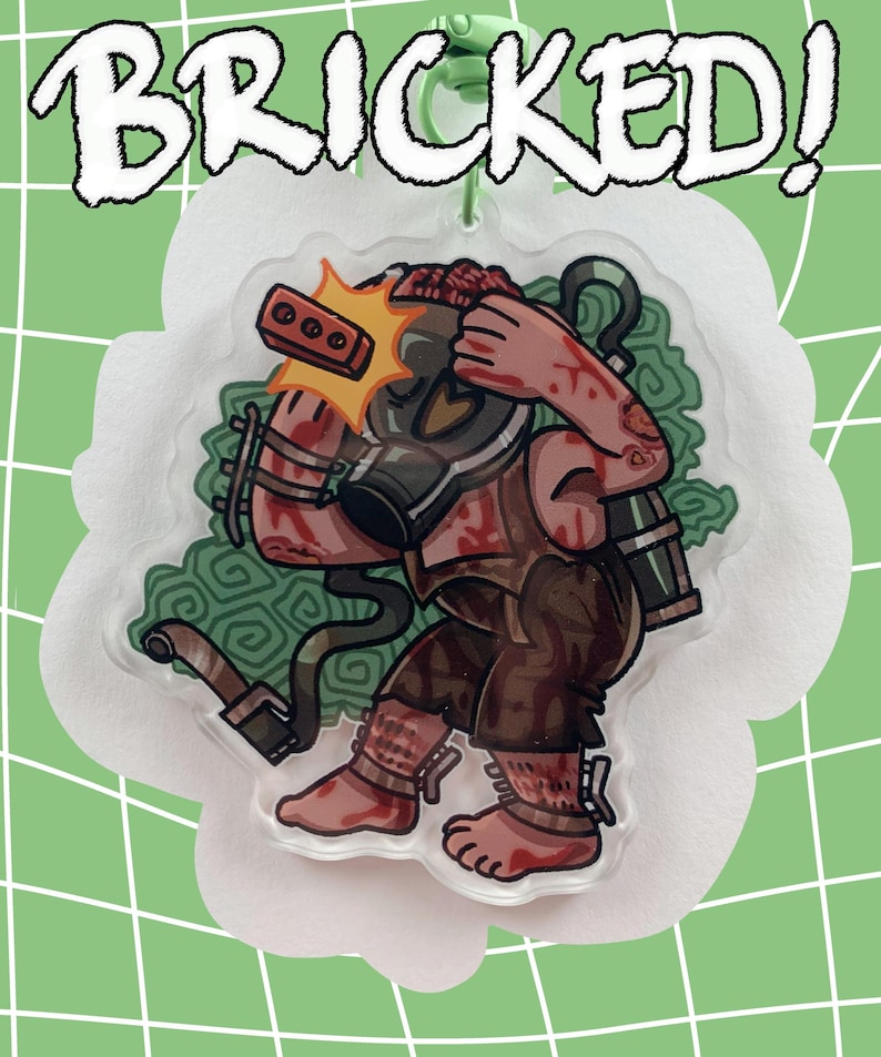 May include: A clear acrylic charm featuring a cartoon figure wearing a gas mask and brown clothing, with blood splatters. The charm has the word "BRICKED!" in white, outlined letters. The charm is on a white, cloud-shaped background, with a green background.