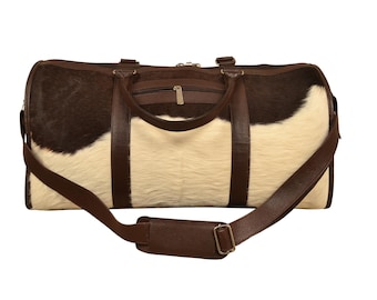 May include: A brown and white cowhide duffel bag with brown leather handles and a shoulder strap. The bag has a zippered top and a small front pocket. The bag is designed for travel and has a rectangular shape.
