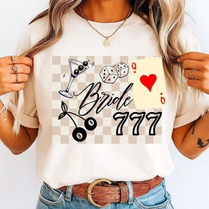 May include: Cream t-shirt with a graphic design including a martini glass, dice, playing card, cherries, and the words "Bride 777". The design has a checkered background.