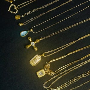 Puede incluir: Una colección de collares dorados con varios diseños de colgantes, incluyendo corazones, cruces y piedras preciosas. Los collares se exhiben sobre una superficie negra, mostrando sus delicadas cadenas y detalles intrincados. Los colgantes presentan una gama de colores y formas.