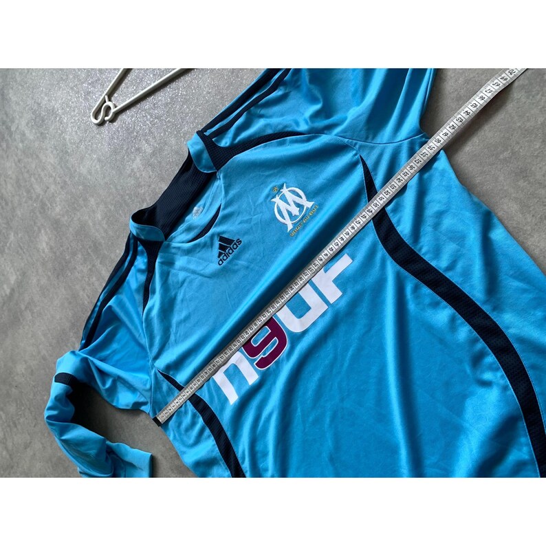 May include: Light blue Adidas soccer jersey with black accents and the Olympique de Marseille logo. The jersey has the Adidas logo and the word "Marseille" in white. A measuring tape is laid across the jersey.